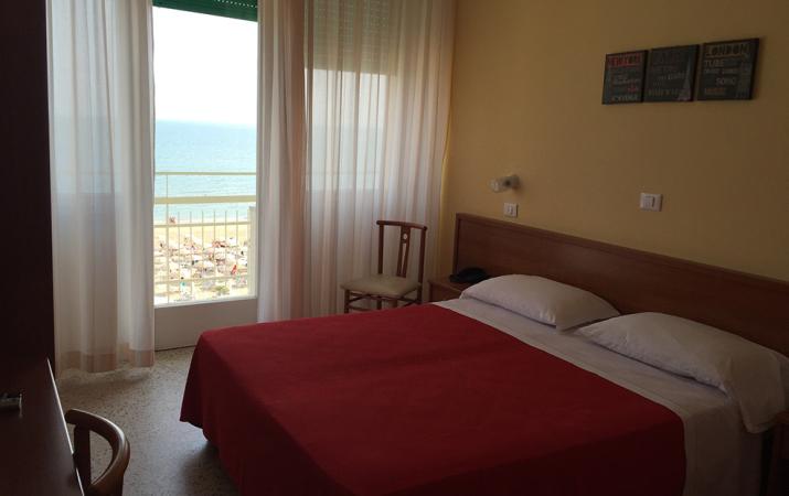 Camera d'albergo con letto matrimoniale, copriletto rosso e balcone con vista.