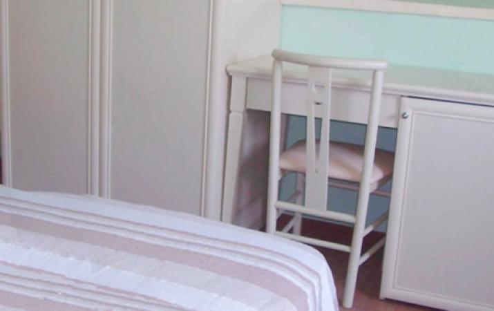 Camera con letto, scrivania, sedia e armadio, arredamento semplice e colori chiari.